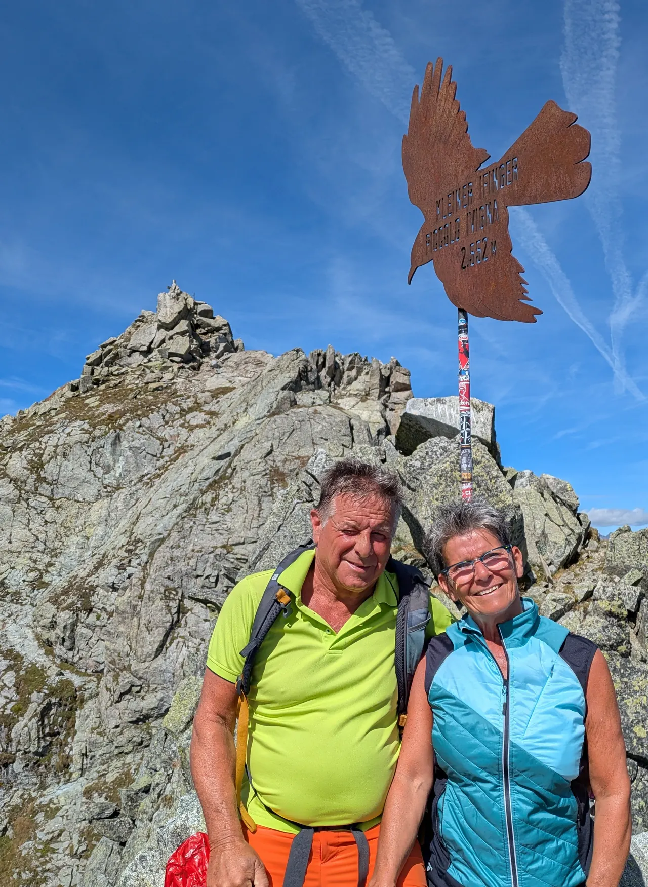 Corina und Donat in Wanderkleidung auf einem Berggipfel vor einem Gipfelkreuz in Form eines Adlers