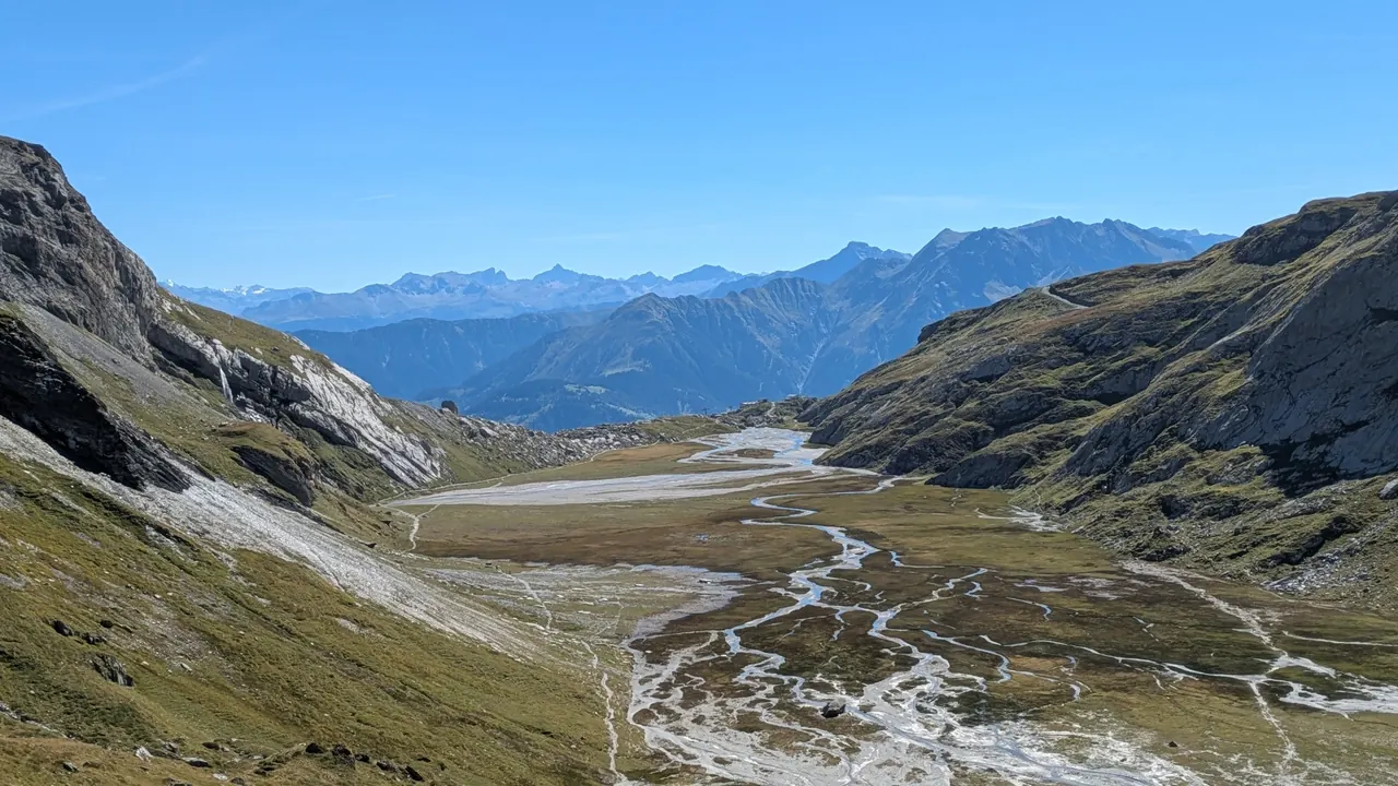 Hochtal mit verzweigten Wasserläufen, dahinter Berge und blauer Himmel