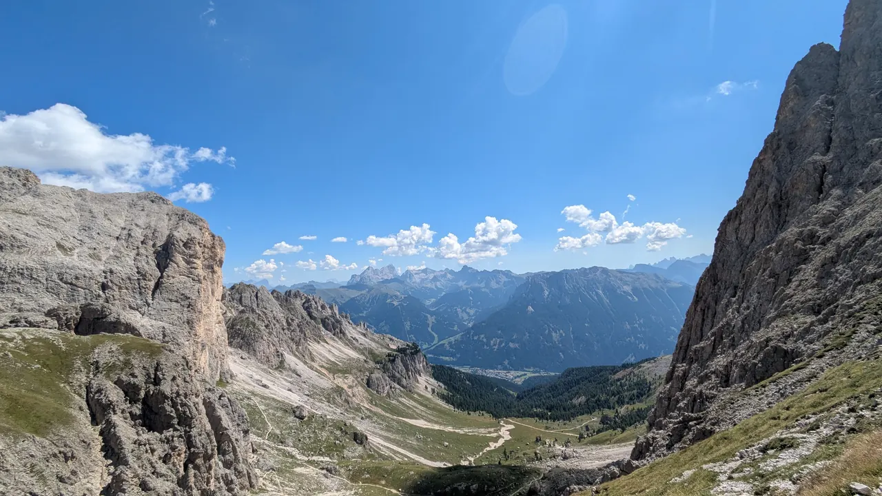 Blick in ein weites Hochtal mit Felswänden, grünen Hängen, fernen Bergketten und blauem Himmel